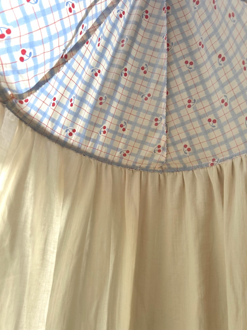 May include: A garment featuring a top section with a light blue and white checkered pattern adorned with small red cherry prints. This section is joined by a horizontal seam to a solid, off-white, gathered fabric skirt that hangs downward.