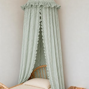 May include: A sage green and white gingham fabric canopy featuring a conical top with a ruffled edge and long, flowing panels that drape downward. The canopy includes a vertical ruffled trim along the front opening, creating a soft, layered look.