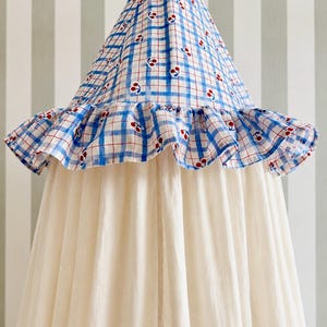 May include: A conical fabric canopy featuring a blue and white checkered pattern with small red cherry motifs. The top section includes a ruffled trim edge, while the lower portion consists of long, plain off-white fabric panels hanging down.