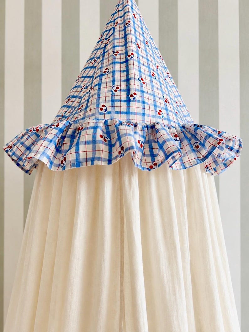 May include: A conical fabric canopy featuring a blue and white checkered pattern with small red cherry motifs. The top section includes a ruffled trim edge, while the lower portion consists of long, plain off-white fabric panels hanging down.