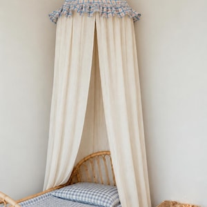May include: A conical bed canopy featuring a blue and white checkered fabric top with a ruffled edge and long, flowing off-white curtains. The canopy is suspended above a rattan bed frame with blue and white checkered bedding and a matching pillow.