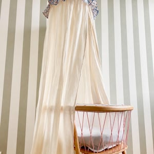 May include: A cream-colored fabric canopy hangs above a wooden bassinet. The canopy features a blue and white checkered top with a ruffled edge. The bassinet has a light wood frame with pink metal wire sides and a white mattress inside.