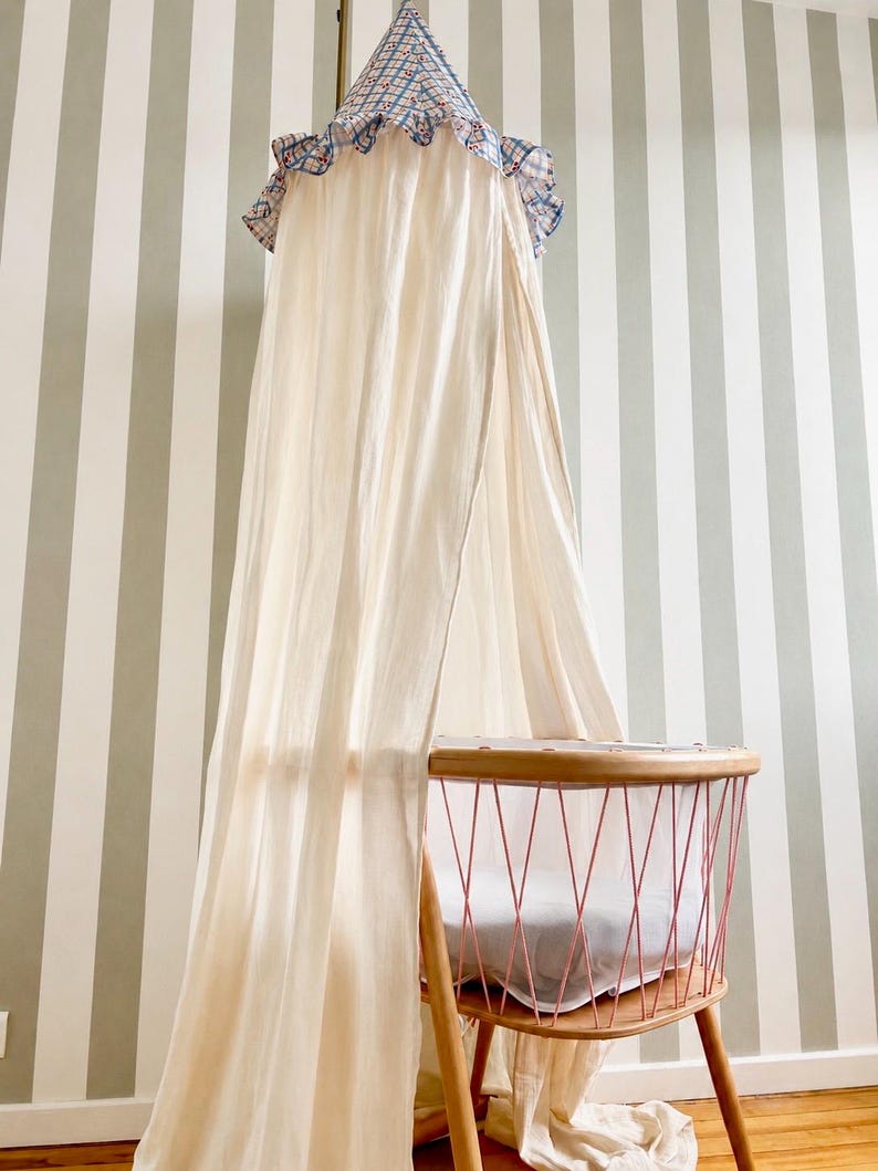 May include: A cream-colored fabric canopy hangs above a wooden bassinet. The canopy features a blue and white checkered top with a ruffled edge. The bassinet has a light wood frame with pink metal wire sides and a white mattress inside.