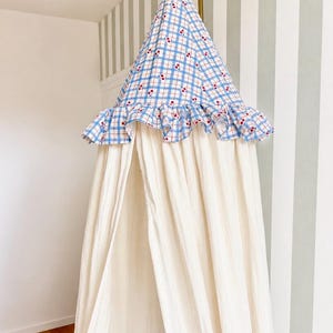 May include: A hanging canopy featuring a conical top made of blue and white checkered fabric with a small red cherry print. The top section includes a ruffled trim, while the lower portion consists of long, solid off-white fabric panels.