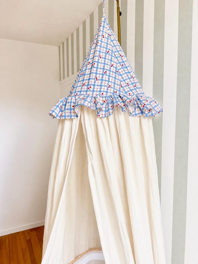 May include: A hanging canopy featuring a conical top made of blue and white checkered fabric with a small red cherry print. The top section includes a ruffled trim, while the lower portion consists of long, solid off-white fabric panels.