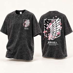 Op de afbeelding: Een antracietgrijs acid-wash T-shirt met het Attack on Titan-logo en roze kersenbloesems. De voorkant heeft een kleine borstafbeelding, terwijl de achterkant een grote versie van hetzelfde ontwerp toont met Japanse tekst en Attack on Titan.