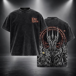 Può includere: Una t-shirt nera con effetto lavaggio acido che presenta il logo The Lord of the Rings sul petto. Il retro mostra una grande grafica dettagliata di una figura in armatura scura con elmo a punte, circondata da testo circolare e un anello d'oro.