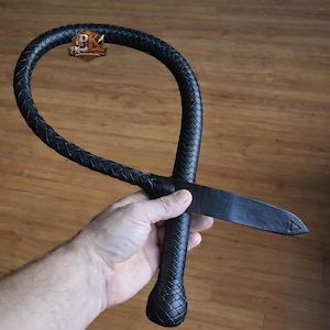 Puede incluir: Látigo de cuero negro con un mango trenzado y un extremo plano y puntiagudo. El látigo se sostiene en una mano, mostrando su artesanía. El logotipo "PKraft Leather" es visible en el fondo.
