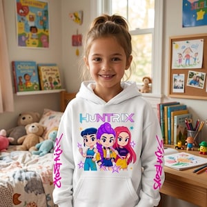 May include: A white hooded sweatshirt featuring the word "HUNTRIX" in blue and purple, with a cartoon graphic of three girls. The sleeves have the text "Girl Boys" in pink. The sweatshirt is worn by a smiling child.