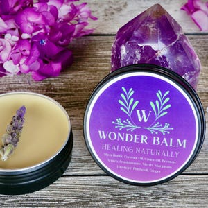 May include: A black tin of Wonder Balm with a purple label and the words "Healing Naturally". A second tin contains a balm with a lavender sprig. Purple flowers and a crystal are in the background.