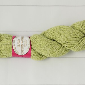 May include: A skein of Plymouth Yarn Llama Cotton Worsted in a light green color. The yarn is twisted and has a label with the yarn name and composition. The yarn is on a white wooden surface.