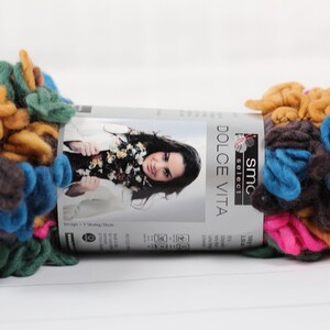 May include: A skein of multicolored yarn with a fuzzy texture. The yarn is in shades of brown, green, blue, pink, and yellow. The yarn is labeled "SMC Select Dolce Vita" and is 100g.
