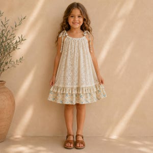 Puede incluir: Un vestido sin mangas de color crema con un intrincado encaje superpuesto en el corpiño y la falda. El dobladillo está acentuado con una hilera de pequeñas borlas y una tela con estampado floral en tonos azules y amarillos.