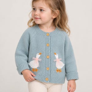 May include: A light blue long-sleeve knit cardigan featuring a crew neckline, ribbed cuffs, and a button-down front with four wooden buttons. Two white goose appliqués with pink tulle accents are stitched onto the lower front panels.