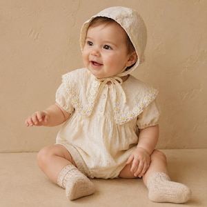 May include: A cream-colored baby romper featuring short puff sleeves, a wide collar with embroidered white and yellow daisy flowers, and a matching bonnet with ties. The outfit is paired with textured knit socks in a coordinating cream shade.