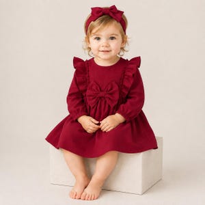 May include: A deep red long-sleeved dress featuring ruffled shoulders, a large central bow on the bodice, and a matching headband with a bow. The garment has a textured fabric finish, long sleeves with gathered cuffs, and a flared skirt design.