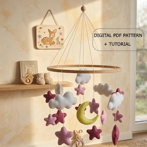 Puede incluir: Móvil para bebé de madera con estrellas, nubes y una luna creciente de fieltro. El móvil está suspendido de un aro de madera. El texto "DIGITAL PDF PATTERN + TUTORIAL" es visible. Un letrero de madera con un ciervo y un conejo está en la pared.