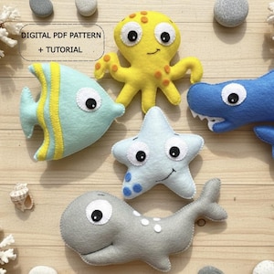May include: A collection of felt sea creatures, including a yellow octopus, blue shark, light blue fish, gray whale, and light blue starfish. The fish has yellow stripes. The text "DIGITAL PDF PATTERN + TUTORIAL" is visible.