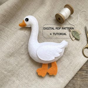 May include: A handmade white felt goose with orange feet and beak. The goose has a black eye and is positioned on a beige fabric. Text on the image reads "DIGITAL PDF PATTERN + TUTORIAL". A wooden spool of thread and scissors are also visible.
