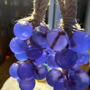 Puede incluir: Un racimo de uvas de cristal translúcido azul-púrpura cuelga de una gruesa cuerda trenzada. Las uvas varían en tonalidad, algunas parecen más oscuras que otras. La cuerda es de color marrón claro natural. El fondo está borroso.