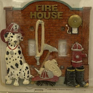 Op de afbeelding: Een rechthoekige lichtschakelaar-afdekplaat met brandweerthema in de originele verpakking. Het ontwerp toont een dalmatiër met een rode helm, een bakstenen muur, een gouden bel, een brandkraan, laarzen, een opgerolde slang en een bijl. Tekst: Switchplate Cover, FIRE HOUSE, A Gift Today, A Treasure Tomorrow.
