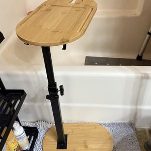 May include: A bamboo bath tray with a black adjustable stand. The tray has a cutout for a wine glass and a rectangular section for a phone or other items. The base is oval-shaped and made of bamboo.