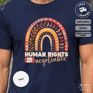 Könnte beinhalten: Marineblaues T-Shirt mit einem farbenfrohen Regenbogen-Design und dem Text "HUMAN RIGHTS ARE NOT negotiable". Der Regenbogen hat orange, gelbe und braune Akzente. Das Shirt besteht aus weicher Baumwolle und ist unisex.