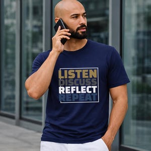 Könnte beinhalten: Marineblaues T-Shirt mit dem Text "LISTEN DISCUSS REFLECT REPEAT" in Weiß, Gold und Blau. Eine Person trägt das T-Shirt und hält ein schwarzes Smartphone an ihr Ohr.