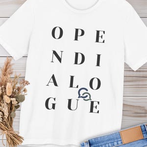 Könnte beinhalten: Weißes T-Shirt mit den Wörtern "OPEN DIALOGUE" in Schwarz vertikal ausgerichtet. Eine blaue Sprechblasen-Grafik ist in das Wort "DIALOGUE" integriert. Das Shirt besteht aus einem weichen Material.