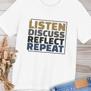 Könnte beinhalten: Weißes T-Shirt mit dem Schriftzug "LISTEN DISCUSS REFLECT REPEAT" in einer schwarzen Box. Der Text ist in Gold, Grau, Schwarz und Marineblau gehalten. Das Shirt liegt auf einer hellen Holzoberfläche mit einem Strauß Trockenblumen und einer Jeans.