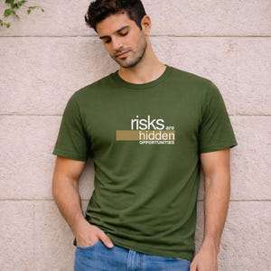 Può includere: T-shirt girocollo verde oliva con la scritta bianca "risks are hidden opportunities". Il testo è sopra un rettangolo marrone. La maglietta è indossata da una persona.