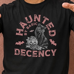 Pode incluir: Uma t-shirt preta com um gráfico de um gato preto a usar óculos redondos ao lado de uma abóbora cheia de doces. O texto HAUNTED BY BASIC DECENCY está escrito em rosa seco, acompanhado por pequenas silhuetas de morcegos e folhas de outono.