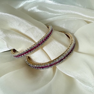 May include: A pair of gold-toned bangles featuring a central row of square-cut deep pink stones flanked by two rows of small, clear, sparkling stones. The circular bracelets have a metallic finish and a structured, multi-layered design.