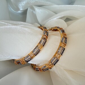 May include: Two circular bangles featuring a gold-toned metal frame with alternating segments of clear, sparkling stones set in silver-toned channels. The design consists of repeating rectangular links that create a structured, segmented appearance.
