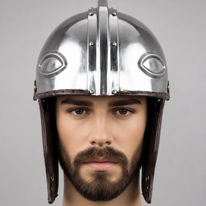 May include: A polished silver-colored metal helmet featuring a central vertical ridge, raised eye-shaped decorative elements on the sides, and brown leather cheek guards attached with visible rivets along the lower edge of the dome.
