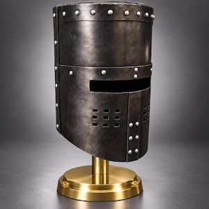 Könnte beinhalten: Ein dunkelgrauer Topfhelm aus Metall in zylindrischer Form mit einem horizontalen Sehschlitz und quadratischen Belüftungslöchern an der Vorderseite. Der Helm ist mit sichtbaren silberfarbenen Nieten versehen und steht auf einem runden goldfarbenen Sockel.