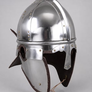 May include: A polished steel helmet featuring a rounded dome, a central ridge, and a T-shaped nasal guard. The helmet includes hinged cheek plates with dark brown leather trim and small brass rivets securing the metal components together.