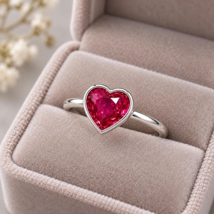 May include: A silver-toned ring featuring a single heart-shaped, faceted red gemstone set in a smooth bezel. The ring is displayed in a neutral-coloured, velvet-lined jewellery box, highlighting the vibrant red colour and the polished metal band.
