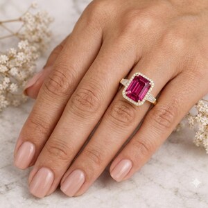 Könnte beinhalten: Ein Goldring mit einem großen, rechteckigen, smaragdgeschliffenen rosa Edelstein als Mittelstein. Der Mittelstein ist von einem Halo aus kleinen, runden, klaren Steinen umgeben, mit zusätzlichen klaren Baguette-Steinen am Goldring.