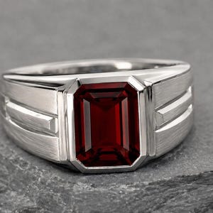 May include: A silver-toned ring featuring a large, rectangular, emerald-cut deep red gemstone set in the center. The band has a brushed metal finish with two horizontal grooved lines on each side of the central stone setting.