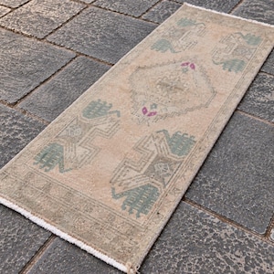 Op de afbeelding: Een rechthoekig loperkleed met een beige basis en geometrische patronen in gedempt saliegroen en accenten in fel magenta. Het kleed heeft een centraal ruitmotief en symmetrische ontwerpen aan beide uiteinden, afgewerkt met korte witte franjes aan de randen.