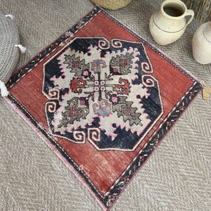 May include: A square rug featuring a central geometric medallion in shades of cream, charcoal, and muted red. The design includes intricate floral-inspired motifs and a dark border with repeating scroll patterns, finished with short fringe edges.