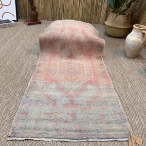 Puede incluir: Una alfombra pasillera rectangular con una paleta de colores desvaídos y suaves en rosa empolvado, azul claro y crema. La alfombra presenta un patrón central de medallón geométrico con detalles desgastados y un borde con flecos.