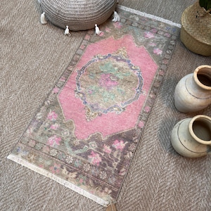 Puede incluir: Una alfombra rectangular con un medallón central rosa rodeado por un borde marrón con patrones florales en tonos rosa, verde y azul. La alfombra presenta una textura desgastada y detalles de flecos blancos en los bordes cortos.