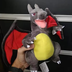 May include: A plush Charizard toy in shades of gray, red, and yellow. The dragon has a yellow belly, red wings, and white claws. The toy is soft and cuddly, with a detailed design.