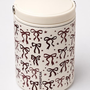 May include: Cream-colored cylindrical container with a matching lid. The container is adorned with a repeating pattern of dark brown bows and stars. The text "HYDRAPEAK" is printed vertically on the side. The lid has a silver clasp.