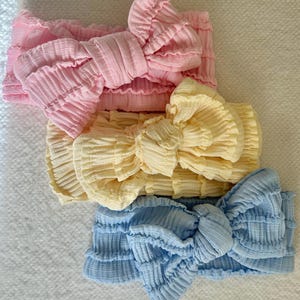 May include: Three pastel-coloured baby headbands in pink, yellow, and blue. Each headband features a large bow in the centre. The fabric has a textured, crinkled appearance.