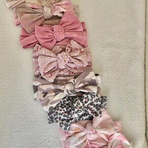 May include: A collection of baby headbands in various shades of pink, beige, and white. The headbands feature different patterns, including floral, leopard print, and checkered designs. The bows are knotted in the center.