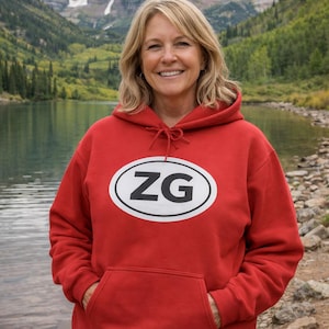 以下が含まれることがあります： 黒い文字「ZG」が入った白い楕円のデザインが施された赤いフード付きスウェットシャツ。スウェットシャツにはフロントポケットとドローストリングフードが付いています。湖と山の前に立っている人。