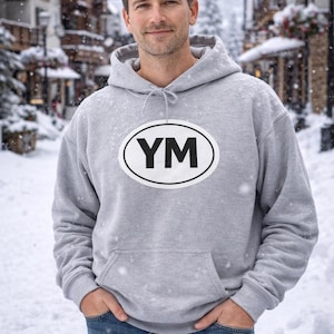 May include: A light gray hoodie with a white oval graphic that has the letters "YM" in black. The hoodie has a drawstring hood and a front pocket. The person is wearing blue jeans and standing in a snowy outdoor setting.